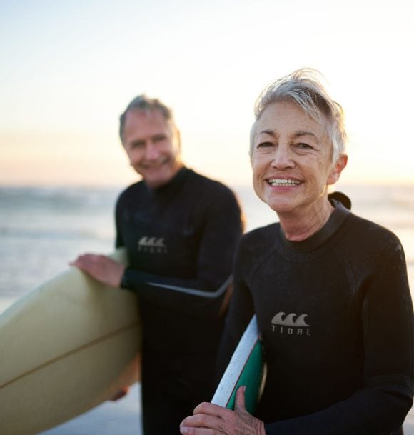 Two elderly couples smiling and holding surfboards while standing in the sea.