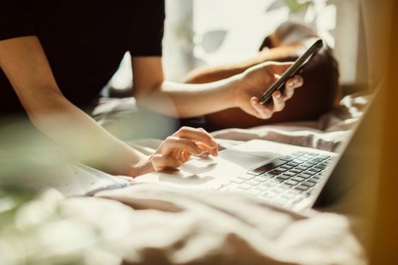pessoa segurando um celular e usando um notebook; ao fundo, um cachorro deitado.
