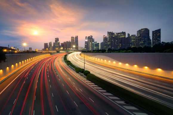 Rodovia urbana à noite com rastros de luz dos carros e prédios iluminados ao fundo