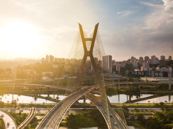 Vista urbana ao pôr do sol com uma ponte em primeiro plano e prédios da cidade ao fundo.
