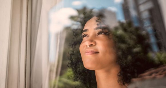 Close no rosto de uma mulher com cabelos cacheados olhando pela janela com expressão contemplativa