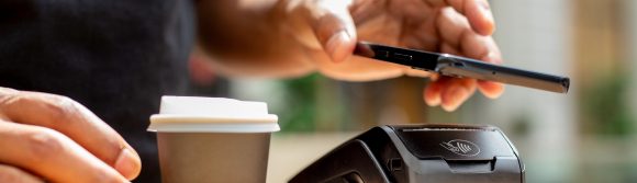 Eine Hand bezahlt einen Take-away-Kaffee mit einem Smartphone über ein Kartenterminal