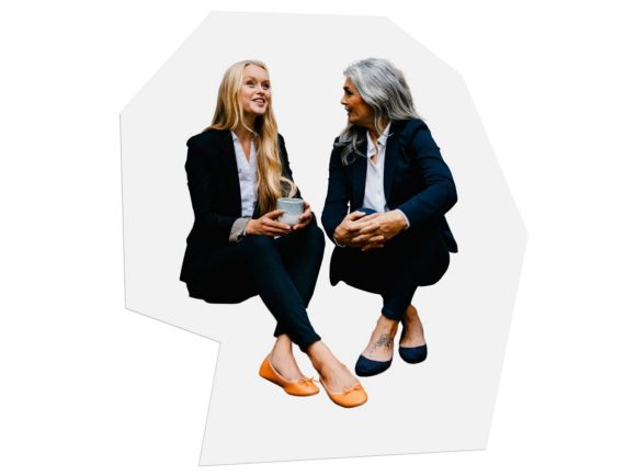 Two women in business attire are sitting next to each other having a conversation.