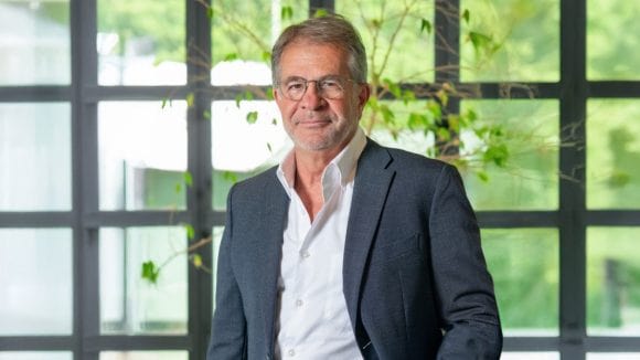 Ein Mann mittleren Alters trägt einen blauen Anzug und blickt offen in die Kamera. Hinter ihm kann man durch eine Glasfront die grüne Aussenfläche sehen.