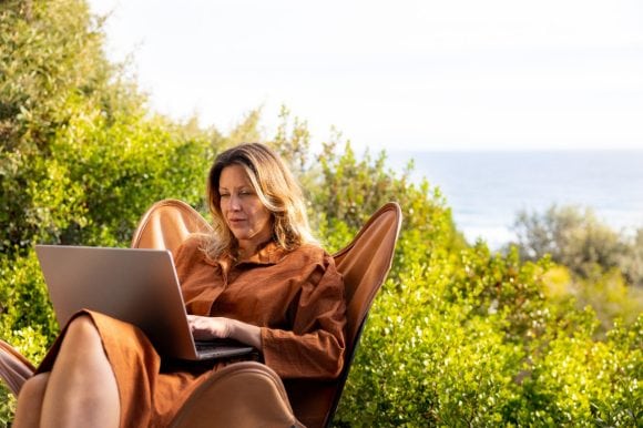 Frau sitzt mit Laptop auf einem Stuhl, hinter ihr Landschaft und Meer