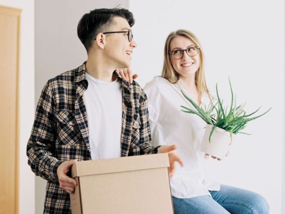 A laughing young couple is holding a moving box and a potted plant.