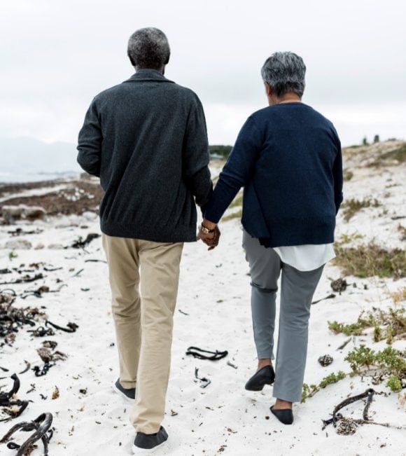 Älteres Paar spaziert Hand in Hand am Strand (Rückenansicht).
