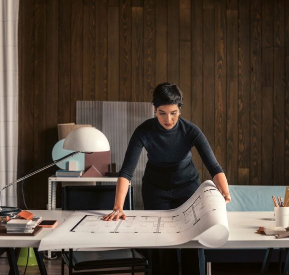 Eine Frau mit kurzen, schwarzen Haaren steht an einem Tisch und überblickt einen grossen, ausgerollten Plan, der einem Bauplan gleicht.