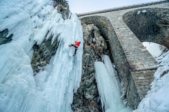 Climber in a red jacket ascending a frozen waterfall beside a stone bridge