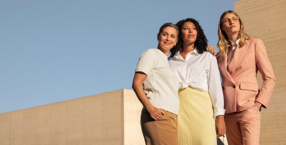 Drei Frauen stehen selbstbewusst vor blauem Himmel und beigefarbenen Betonwänden