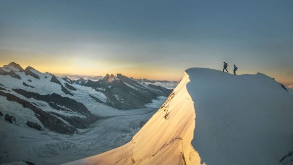 Two hikers stand on a mountain summit surrounded by mountains.