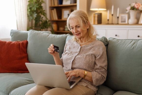 An older woman sits comfortably on her sofa, using a laptop and credit card to make a secure online purchase
