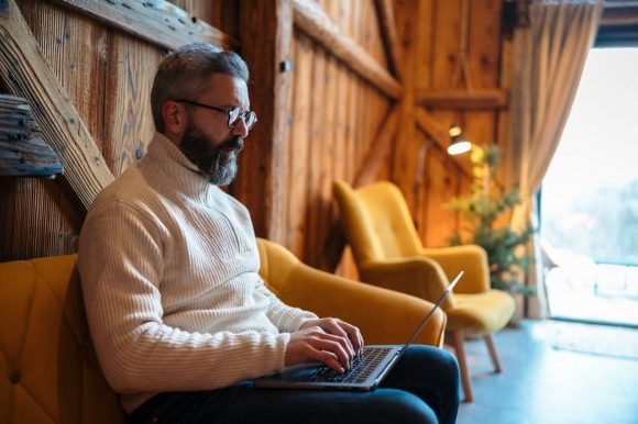 Bearded man in a beige zip neck jumper sitting on an ochre sofa with a laptop