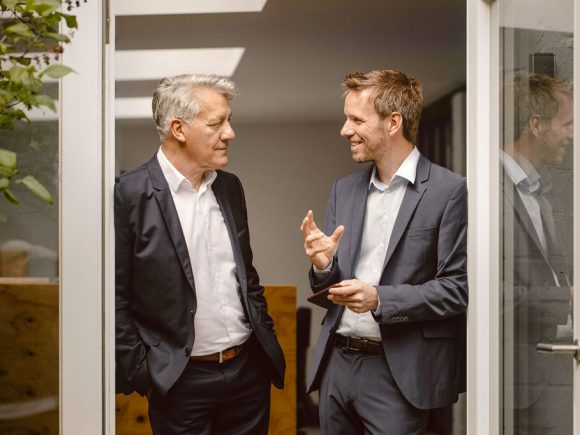 Two men in suits are talking at a glass door.