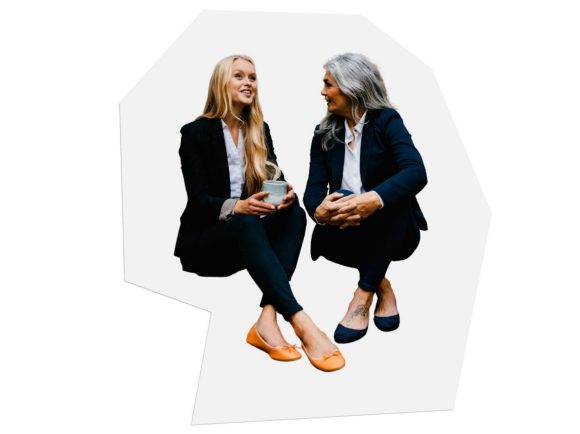 Two women in business attire are sitting next to each other having a conversation.