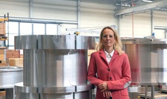 A woman with blonde hair and a red blazer stands smiling in front of large metal tubes in an industrial hall.