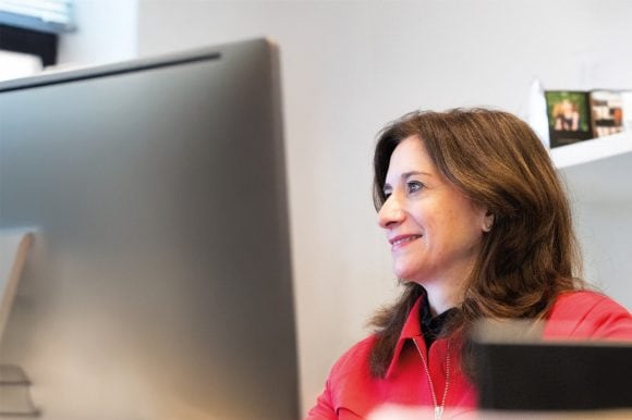 A woman with long brown hair and a red top sits at a desktop computer and smiles contentedly.