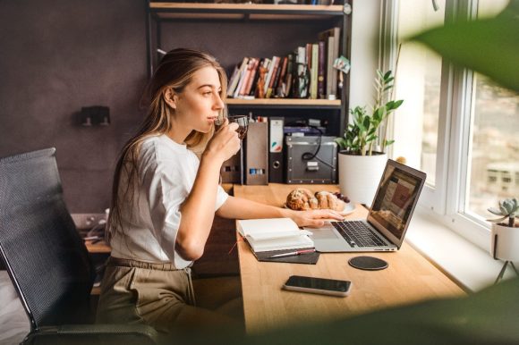 A young woman sits at her desk in front of her laptop drinking tea and looking out the window.