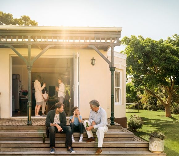 Three people are sitting relaxed on the terrace of a house, drinking together. In the background, three women are inside the house, chatting and also drinking – the mood is cheerful and festive after the successful house purchase.