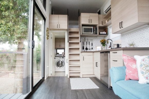 Modern micro-apartment with kitchen, living area and loft bed in a small space; floor-to-ceiling windows offer a view of the outdoors.
