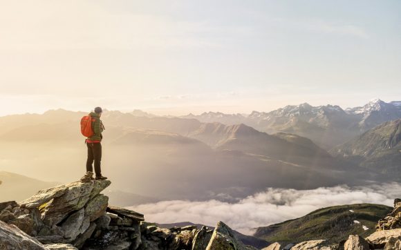 A hiker is standing on the summit of a mountain looking out over the mountain range towering above the clouds.