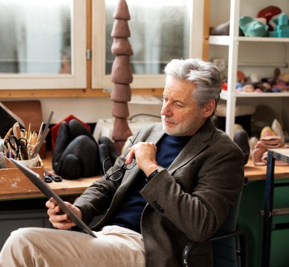 A man with gray hair and beard sits on a chair in a studio examining a document while holding his glasses in his left hand.