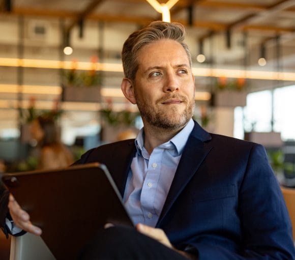 A blond middle-aged man in a suit sits on a chair with a tablet in his hand and looks into the distance.