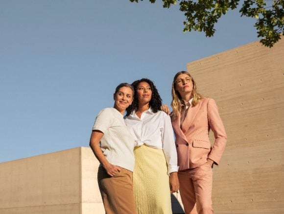 Three women standing confidently against a blue sky and beige concrete walls