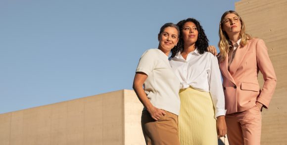 Three women standing confidently against a blue sky and beige concrete walls