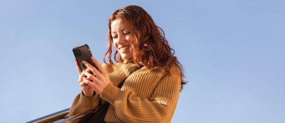 Femme rousse en pull ocre souriant à un smartphone