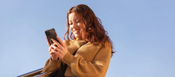 Femme rousse en pull ocre souriant à un smartphone