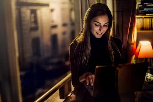 A young woman sits in a wicker chair with her laptop on her lap, holding her hands to her face in shock – cyber fraud alert!
