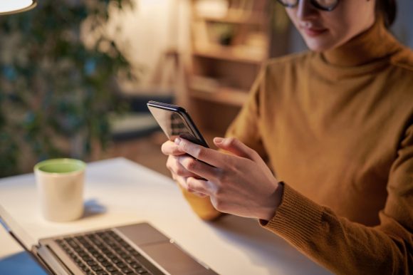 Une jeune femme est assise à la table avec un ordinateur portable et lit un message sur son téléphone.