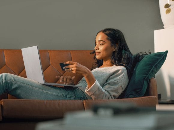 A young woman lying on the sofa using her tablet while holding her UBS Credit Card in her hand.