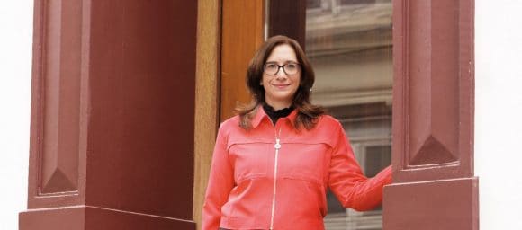 Une femme aux cheveux bruns portant des lunettes et un haut rouge avec une fermeture éclair blanche se tient devant une porte, sa main gauche contre le mur.