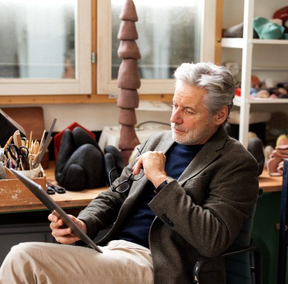 A man with gray hair and beard sits on a chair in a studio examining a document while holding his glasses in his left hand.