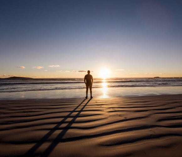 Two men in casual shirts smiling and walking arm in arm on the beach.