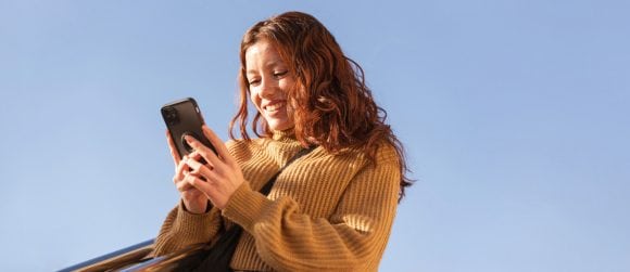 Donna dai capelli rossi con maglione ocra che sorride davanti a uno smartphone