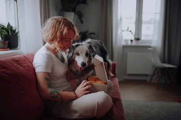 Una donna apre un pacco su un divano con vicino un cane pastore australiano in un accogliente soggiorno.