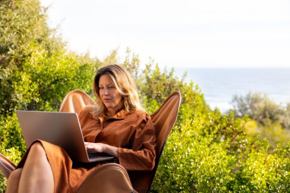 Una donna con un laptop su una poltrona, con vegetazione e mare sullo sfondo