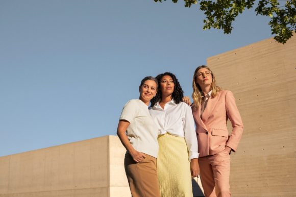 Tre donne stanno in posa con sicurezza davanti a un cielo blu e a pareti in cemento beige