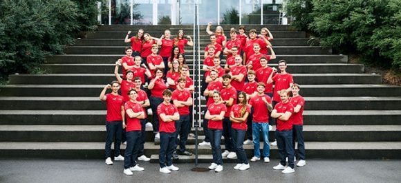 The image shows all 42 young professional talents wearing red jackets, standing together on a wooden boardwalk.