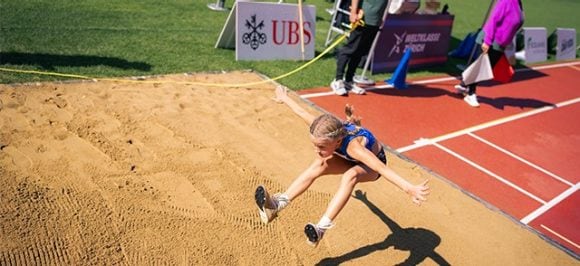 The image shows the long jump discipline at the UBS Kids Cup. A girl is performing a long jump into the sand in a stadium. In the background, you can see the UBS logo and the officials (Kampfrichter).