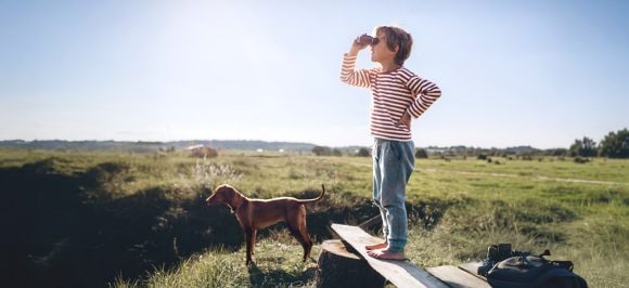 The picture shows a boy standing barefoot on a wooden bench in a green setting, looking into the distance through a telescope.