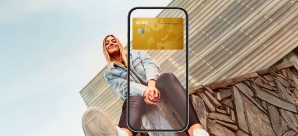 L’image montre une femme assise sur un mur. Au-dessus, un smartphone transparent est superposé, avec une carte de crédit Gold visible dans le portefeuille numérique (Wallet).