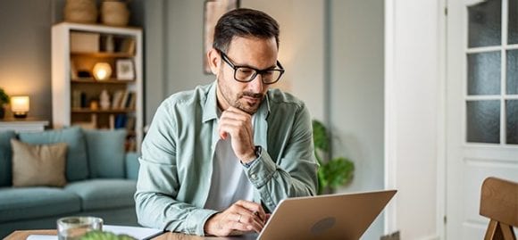Sur cette image, on voit un jeune homme assis à une table dans un salon, regardant attentivement un ordinateur portable.