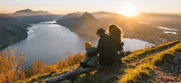 Un paesaggio autunnale con un grande lago e montagne sullo sfondo. In primo piano una famiglia (due adulti e un bambino) è seduta su un tronco d’albero in un punto panoramico.