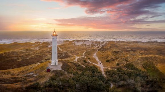 Weisser Leuchtturm inmitten von Sanddünen mit Blick auf einen Sonnenuntergang über dem Meer