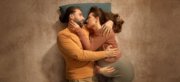 Das Bild zeigt ein liegendes Paar, das sich umarmt. Die Frau ist schwanger und sie freuen sich auf die Zukunft zu Dritt.