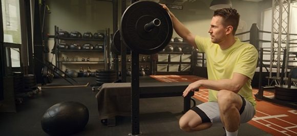 The picture shows a man in a gym, kneeling in front of a piece of equipment and considering how much weight to choose.